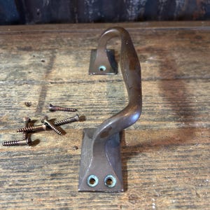 May include: A bronze-colored, curved handle with two rectangular mounting plates. Several bronze-colored screws are scattered nearby. The handle is likely for a cabinet or drawer, and is set against a weathered wooden surface.