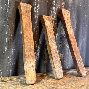 May include: Three rusty metal rods, likely used for construction or blacksmithing, are leaning against a dark, textured background. The rods are weathered and have a rough, uneven surface.
