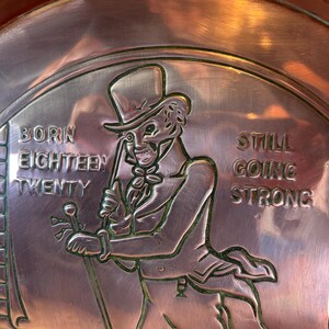 Puede incluir: Un plato color cobre con un dise&ntilde;o en relieve. La figura central es un hombre con sombrero de copa y traje, sosteniendo un bast&oacute;n. El texto dice "BORN EIGHTEEN TWENTY" y "STILL GOING STRONG". El plato tiene un borde decorativo.