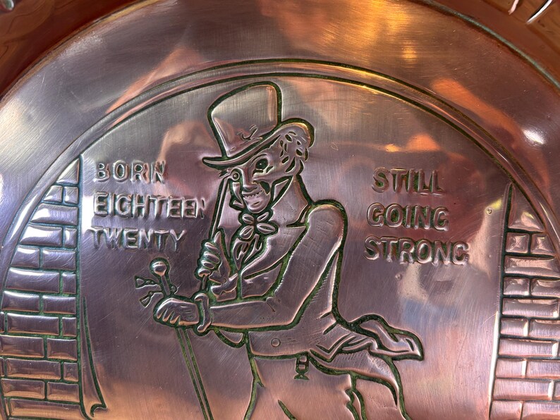 Puede incluir: Un plato color cobre con un dise&ntilde;o en relieve. La figura central es un hombre con sombrero de copa y traje, sosteniendo un bast&oacute;n. El texto dice "BORN EIGHTEEN TWENTY" y "STILL GOING STRONG". El plato tiene un borde decorativo.