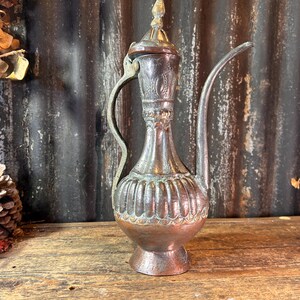 May include: An antique copper-toned metal pitcher with a long spout and ornate detailing. The pitcher has a handle and a decorative lid. The background features a rustic corrugated metal surface and dried flowers.