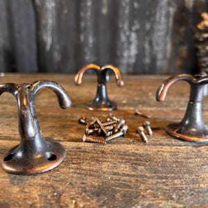 May include: Three antique-style hooks with a dark bronze finish. Each hook has three prongs and a round base. The hooks are arranged on a wooden surface with screws scattered around them.