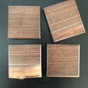 Puede incluir: Cuatro posavasos cuadrados de madera de nogal. Los posavasos son de color marrón oscuro y tienen un patrón de veta de madera natural.