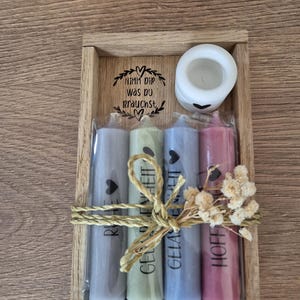 Puede incluir: Una bandeja de madera con cuatro velas, cada una de un color diferente y con una palabra impresa en ella. Las palabras son "Ruhe", "Gelassenheit", "Gelassenheit" y "Hoffnung". La bandeja también tiene un pequeño portavelas blanco y un letrero que dice "Nimm dir was du brauchst".