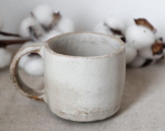 May include: A white ceramic mug with a rounded shape and a brown handle. The mug has a rustic, handmade look.