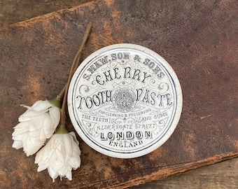 May include: A vintage round tin of Cherry Tooth Paste, with black text and ornate designs on a white background. The text reads "S. Maw, Son & Sons" and "London England". Two white fabric flowers rest on a brown leather surface.