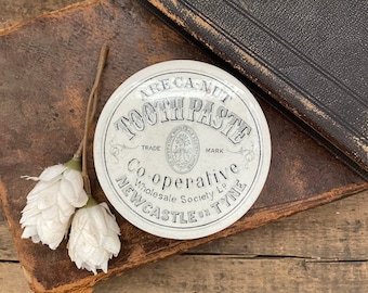 May include: A vintage, round tin of "ARECA-NUT TOOTHPASTE" with text and a logo, resting on a worn, brown leather-bound book. Two white floral sprigs are next to the tin. A dark brown book is in the background.