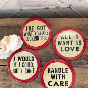 May include: Four round, vintage-style signs with red borders and cream-colored centers. The signs feature black text: "I'VE GOT WHAT YOU ARE LOOKING FOR," "ALL I WANT IS LOVE," "I WOULD IF I COULD, BUT I CAN'T," and "HANDLE WITH CARE."
