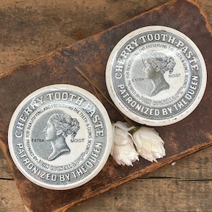 May include: Two vintage white ceramic lids for cherry tooth paste jars. The lids feature a black and white image of a woman's profile with the text "Cherry Tooth Paste" and "Patronized by the Queen".