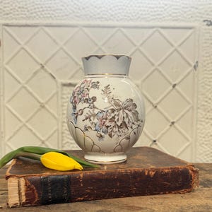 May include: A vintage, round vase with a light blue rim and gold trim, adorned with floral patterns in pink, blue, and brown. The vase sits atop a worn, leather-bound book, with a yellow tulip and green stem resting on the book.