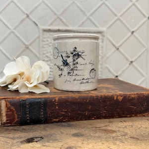 May include: A small, off-white ceramic jar with a vintage design featuring birds and script. The jar has a cylindrical shape and is decorated with blue and black ink illustrations. The text "CARTE POSTALE" is visible on the jar.