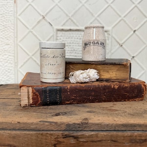 May include: Two antique ceramic jars, one labeled "Sugar" and the other with French text, sit atop aged books. A bundle of dried white flowers rests between the jars. The jars and books are on a wooden surface.