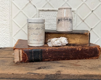 May include: Two antique ceramic jars, one labeled "Sugar" and the other with French text, sit atop aged books. A bundle of dried white flowers rests between the jars. The jars and books are on a wooden surface.
