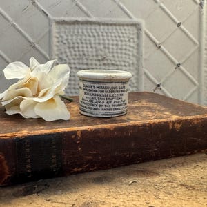 May include: A small white ceramic jar with a black label that reads "Clarke's Miraculous Salve" and lists uses for the salve, including "Ulcerated Bad Legs, Boils, Abscesses, Eczema and All Skin Eruptions". The jar is sitting on top of a brown leather-bound book.