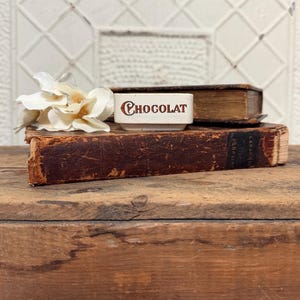 May include: A small, rectangular, off-white ceramic dish featuring the word CHOCOLAT printed in brown lettering on its front face. The dish is positioned next to a cluster of white artificial flowers and rests atop two weathered, brown leather-bound books.