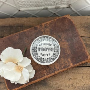 May include: A white ceramic lid with black lettering that reads "Carbolic Tooth Paste". The lid has a decorative border with a floral design.