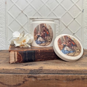 May include: A vintage ceramic jar with a lid, both featuring a colorful illustration of a couple in a historical setting. The jar is white with a round shape and a decorative design. The lid is white with a similar design. The jar is sitting on top of a book.