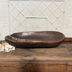 May include: A dark brown, oval wooden bowl with a rustic, aged appearance. The bowl has a shallow depth and a smooth interior. Small, clear decorative accents are placed along the rim. The bowl is displayed on a wooden surface.