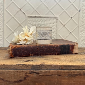May include: A vintage, cylindrical tin of Clarke's Miraculous Salve, with printed text, sits atop a worn, leather-bound book. A cream-colored flower rests beside the tin. The items are on a wooden surface, with a white, patterned background.