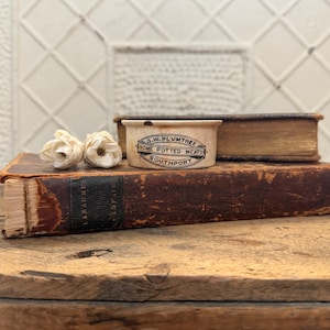 May include: An antique ceramic pot with the text "G.W. Plumtree Home Potted Meats Southport" sits atop a stack of old books. Two cream-colored paper flowers are in the foreground. The books and pot are on a wooden surface.