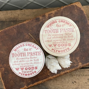 May include: Two vintage round white tins with red lettering. The tins are labeled "Woods Cherry Od Tooth Paste" and contain instructions for removing tartar and whitening teeth without injuring the enamel. The text also includes the proprietor's name, W. Woods, and his profession, Chemist, along with his location, Plymouth.