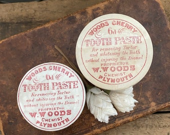 May include: Two vintage round white tins with red lettering. The tins are labeled "Woods Cherry Od Tooth Paste" and contain instructions for removing tartar and whitening teeth without injuring the enamel. The text also includes the proprietor's name, W. Woods, and his profession, Chemist, along with his location, Plymouth.