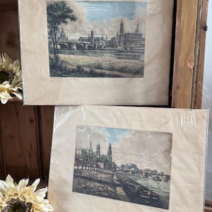 May include: Two framed vintage prints of a European city, likely Dresden, Germany, featuring a river, bridges, and detailed architectural structures. The prints are in protective plastic, with a beige paper backing. The artwork has a muted color palette with hints of blue and green.