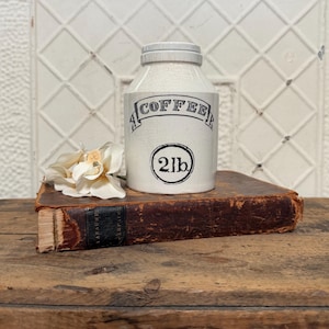 May include: A cream-colored ceramic canister with a crackled glaze finish, featuring the black text COFFEE inside a banner and 2lb inside a circle. The canister has a matching lid and is positioned next to a white flower on a brown book.