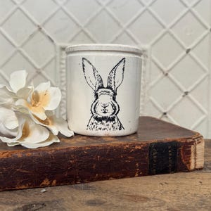 May include: A cream-colored ceramic jar with a black illustration of a rabbit's face. The jar is cylindrical and sits on a vintage book with a dark brown cover. A white flower is to the left of the jar.