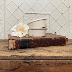 May include: Two stacked off-white ceramic ramekins with a smooth finish, positioned next to a white artificial flower on top of a weathered, dark brown antique book with a black spine label that reads Labaume's Campaigns.
