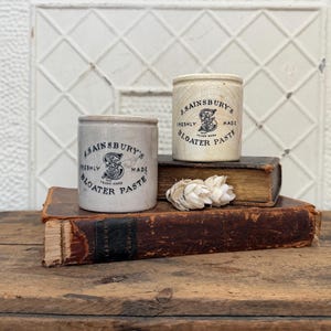 May include: Two antique J. Sainsbury's Bloater Paste jars with black text and a logo. One jar is light beige, the other is off-white. They sit on old books with a white, textured background. The jars are cylindrical with a lid.