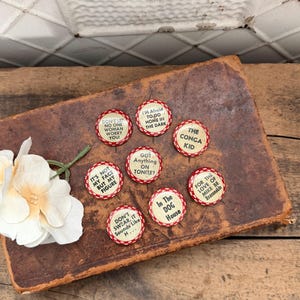 May include: A selection of vintage circular buttons with red and white edging, each bearing a unique phrase. The buttons are displayed on a well-worn, brown leather-bound book. A white flower with a green stem rests on the book.