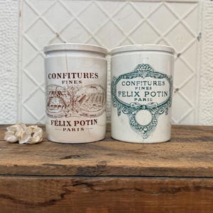 May include: Two antique ceramic jars with lids. One jar has a brown illustration and text that reads "CONFITURES FINES FÉLIX POTIN PARIS". The other jar has a teal illustration and text that reads "CONFITURES FINES FÉLIX POTIN PARIS".
