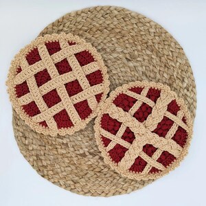 May include: Two crocheted pie-shaped pot holders in red and beige on a woven jute background.