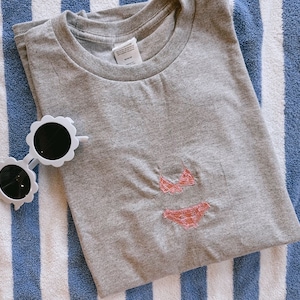 May include: A grey t-shirt with a pink embroidered bikini design on a blue and white striped towel.