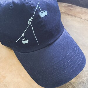 May include: A dark blue baseball cap with a white embroidered design of a ski lift with two gondolas.