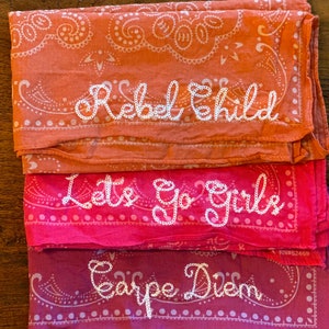 May include: Three pink and orange bandanas with white embroidered text. The top bandana says "Rebel Child", the middle bandana says "Lets Go Girls", and the bottom bandana says "Carpe Diem".