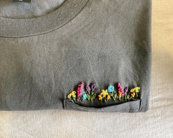 May include: A dark gray t-shirt with a pocket on the left side. The pocket is embroidered with a row of colorful flowers.