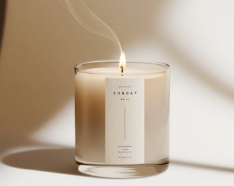 May include: A beige candle in a glass jar with a label that reads "The Scents of SUNDAY NO. 01 100% NATURAL SOY WAX 9 OZ". The candle is lit and smoke is rising from the flame.