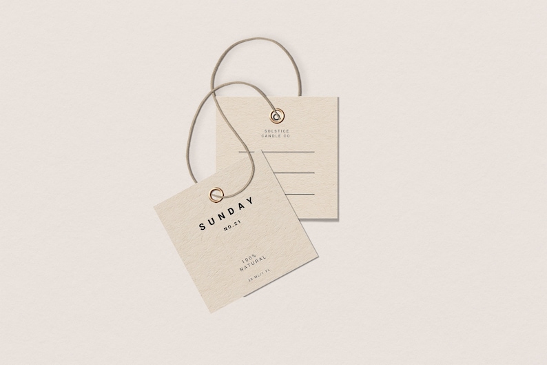 May include: A beige square tag with the text "SUNDAY NO. 21 100% NATURAL 20 ML/FL" and a gold metal ring. A smaller beige square tag with the text "SOLSTICE CANDLE CO." is attached to the larger tag with a brown string.