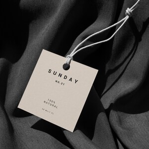 May include: A beige square tag with the text "SUNDAY NO.21 100% NATURAL 30 ML/1 FL" on a dark gray fabric background.