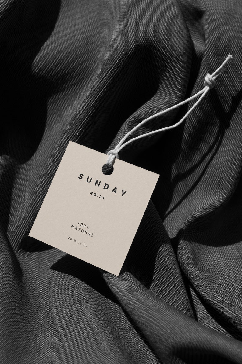 May include: A beige square tag with the text "SUNDAY NO.21 100% NATURAL 30 ML/1 FL" on a dark gray fabric background.