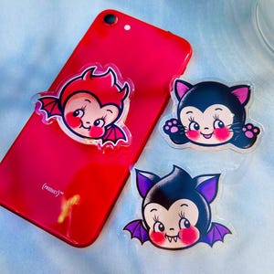 May include: Three clear acrylic phone grips with cartoon bat and cat designs. The bat designs are red and purple, and the cat design is black and pink. One grip is attached to a red phone.