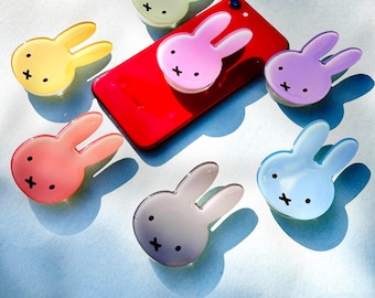 May include: A red smartphone with a pink bunny-shaped phone grip attached, surrounded by six colorful bunny-shaped phone grips in yellow, pink, purple, light blue, peach, and gray. Each bunny has a black cross and dot face.