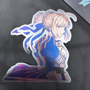 Puede incluir: Una pegatina holográfica con un personaje de anime de cabello rubio, ojos azules y un atuendo azul y plateado. El personaje mira hacia la derecha y tiene una expresión seria.