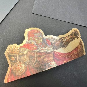 May include: A sticker featuring a detailed illustration of a character with red hair, a red cape, and ornate armor. The sticker has a vintage, slightly aged appearance, with a tan background and a detailed design. The character is posed with one arm outstretched.