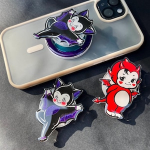 May include: A clear phone case with a pop-up phone grip featuring a cartoon vampire bat design in black, purple, and gray. Two additional matching bat and devil-themed phone grips are also visible, all with a glossy finish.