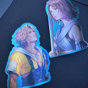 May include: Two die-cut stickers featuring anime-style characters. One sticker depicts a male character with blond hair, a yellow jacket, and blue overalls. The other shows a female character with brown hair and a white and blue outfit.