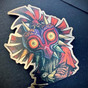 May include: A sticker featuring the Majora's Mask from The Legend of Zelda. The mask is primarily red, purple, and gold, with sharp details and a menacing expression. The sticker has a gold border and is on a black surface.
