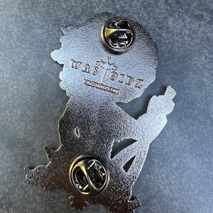 May include: A silver-colored enamel pin with a cartoon figure giving the middle finger. The pin has two pin backs and the text "WAS TOIDZ" and "WARTOIDZ.COM" is visible.